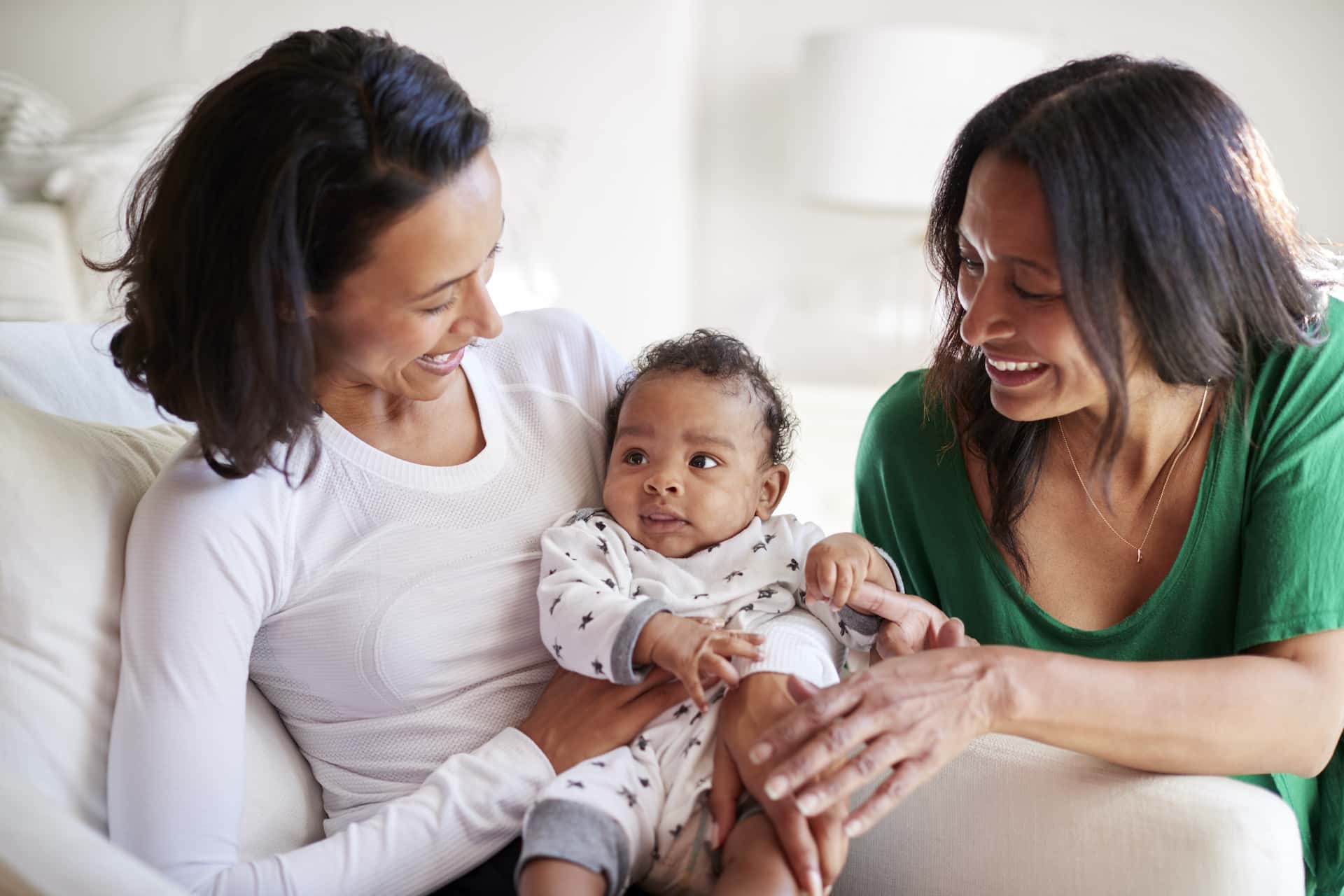 Millennial mixed race mother sitting in an armchair holding her three month old baby son, her mother kneeling beside them Birth Tourism